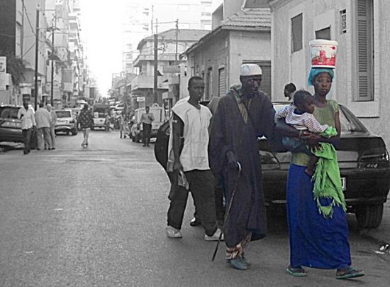 La rue à Dakar, une jeune femme, bébé dans les bras et charge sur la tête guide un vieil aveugle, l'air désabusée. Par Tili